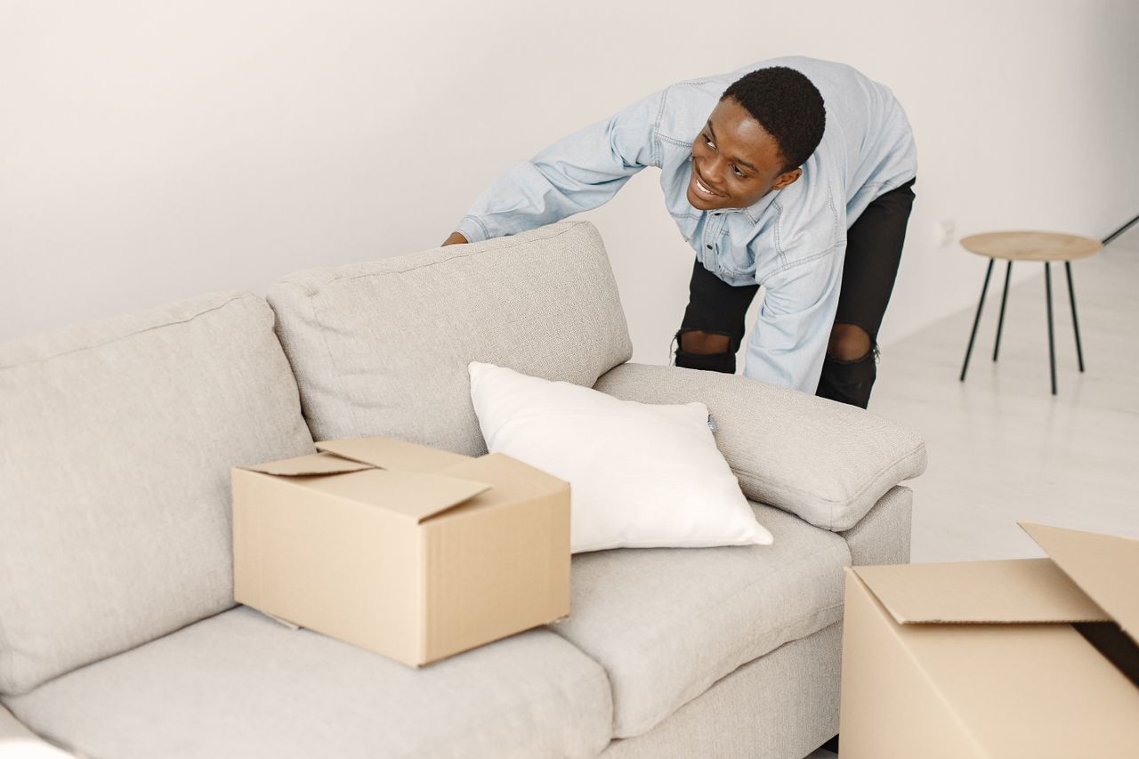 why-choose-us-02 Young man smiling while arranging cardboard boxes in a modern living room.